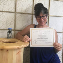 Adina kneels on the floor holding a certificate of certification for Vaginal Steam Facilitator. She is leaning on a round steam stool, in front of a fabric backed grid window casing.